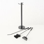 Kaminbesteck Mika, 4-teilig, bestehend aus einem Ständer und drei Werkzeugen, auf einer weißen Oberfläche vor einer weißen Ziegelwand, seitliche Perspektive.