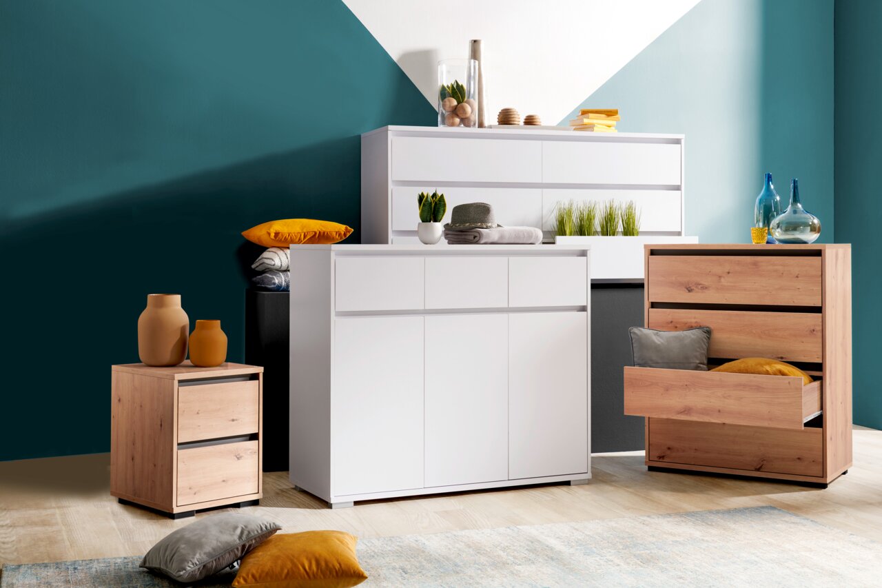 Carryhome Nachtkommode LINE Moderne Kommoden in verschiedenen Größen und Farben, frontal fotografiert. Eine weiße Kommode mit Schubladen und Türen, daneben eine Holzkommode mit offenen Schubladen. Dekorative Elemente wie Vasen und Kissen sind auf und um die Möbel platziert.