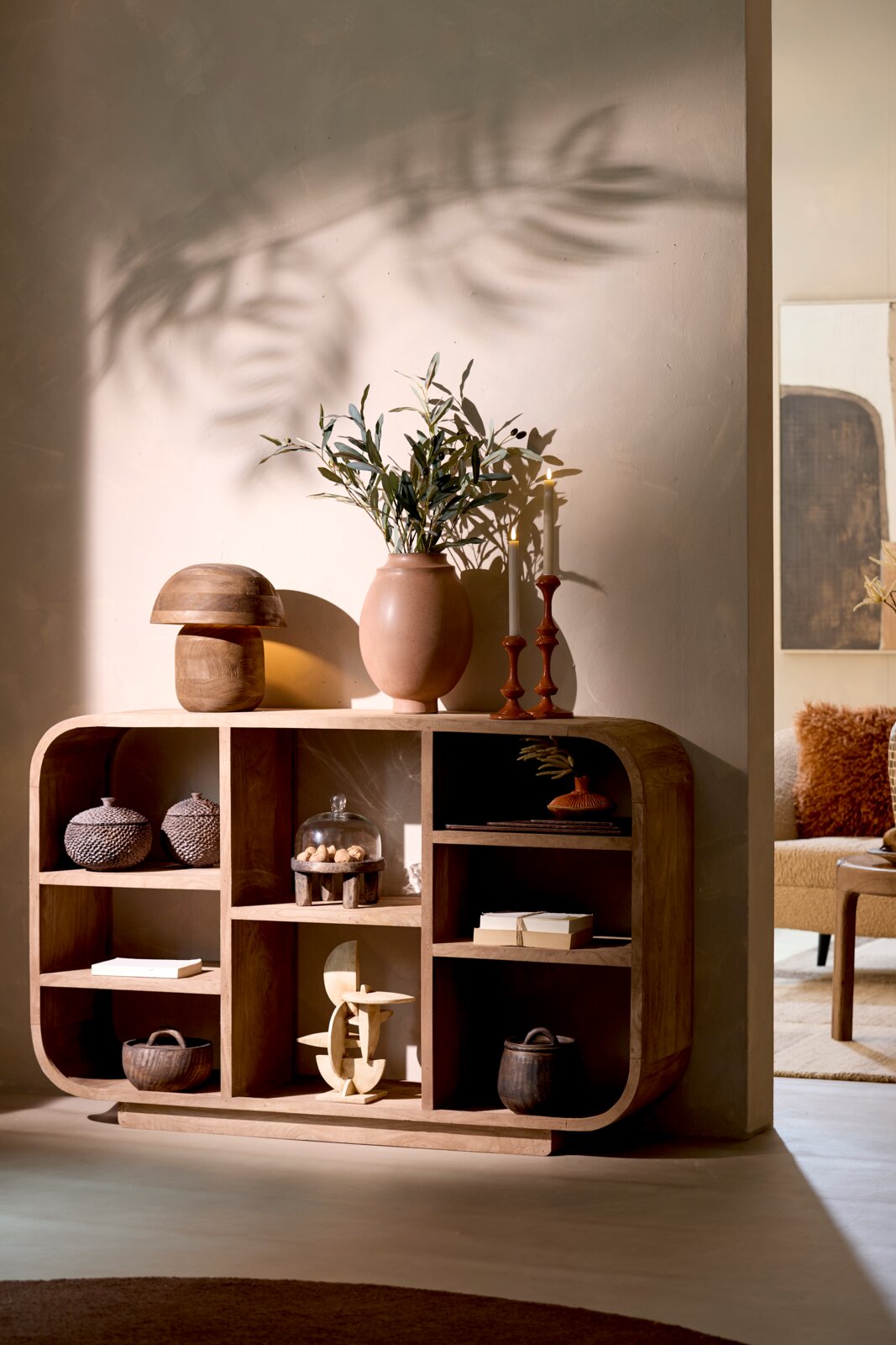 Handgemaakte houten vakkenkast met organisch afgeronde hoeken en asymmetrische open compartimenten, vanuit een licht schuin vooraanzicht sfeervol gestyled met woonaccessoires