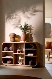 Handgemaakte houten vakkenkast met organisch afgeronde hoeken en asymmetrische open compartimenten, vanuit een licht schuin vooraanzicht sfeervol gestyled met woonaccessoires