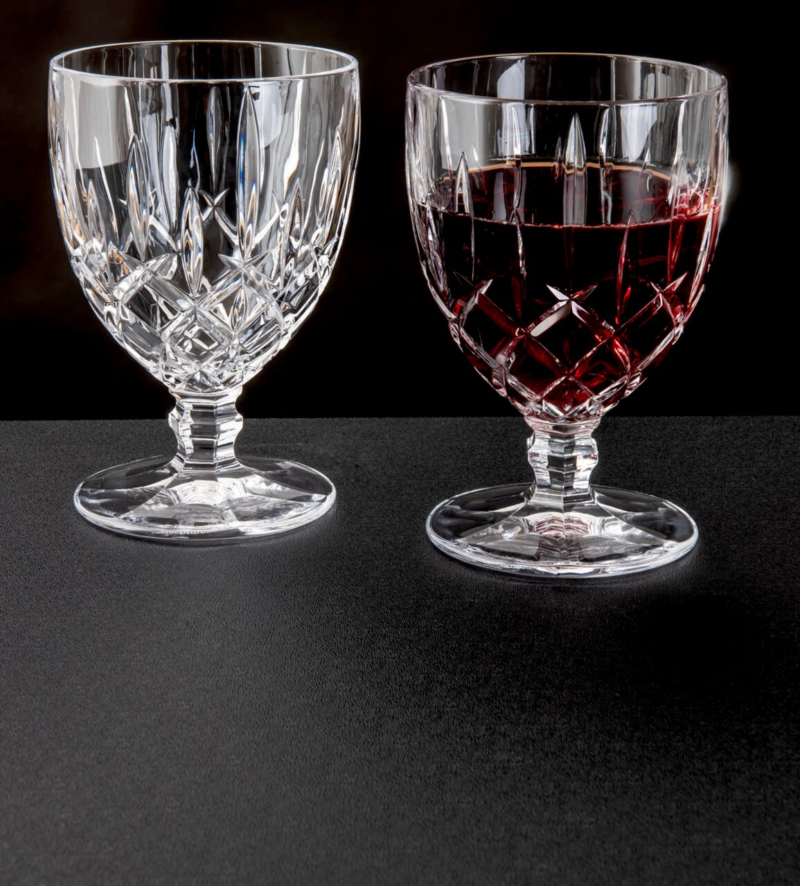 Zwei Weinkelche Noblesse 350ml aus Kristallglas, einer leer und einer mit Rotwein gefüllt, auf schwarzem Untergrund, seitliche Perspektive.