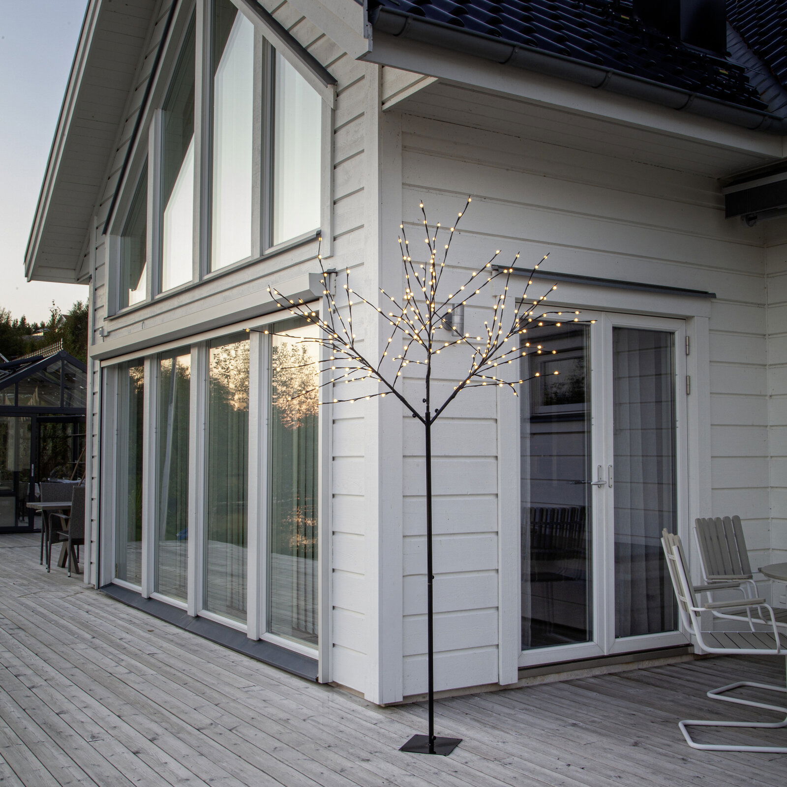 Schwarzer LED-Lichtbaum als dekorative Stehleuchte auf einer Holzterrasse vor einem weißen Haus, warmweiße Lichter, Perspektive: leicht seitliche Außenansicht auf Augenhöhe.