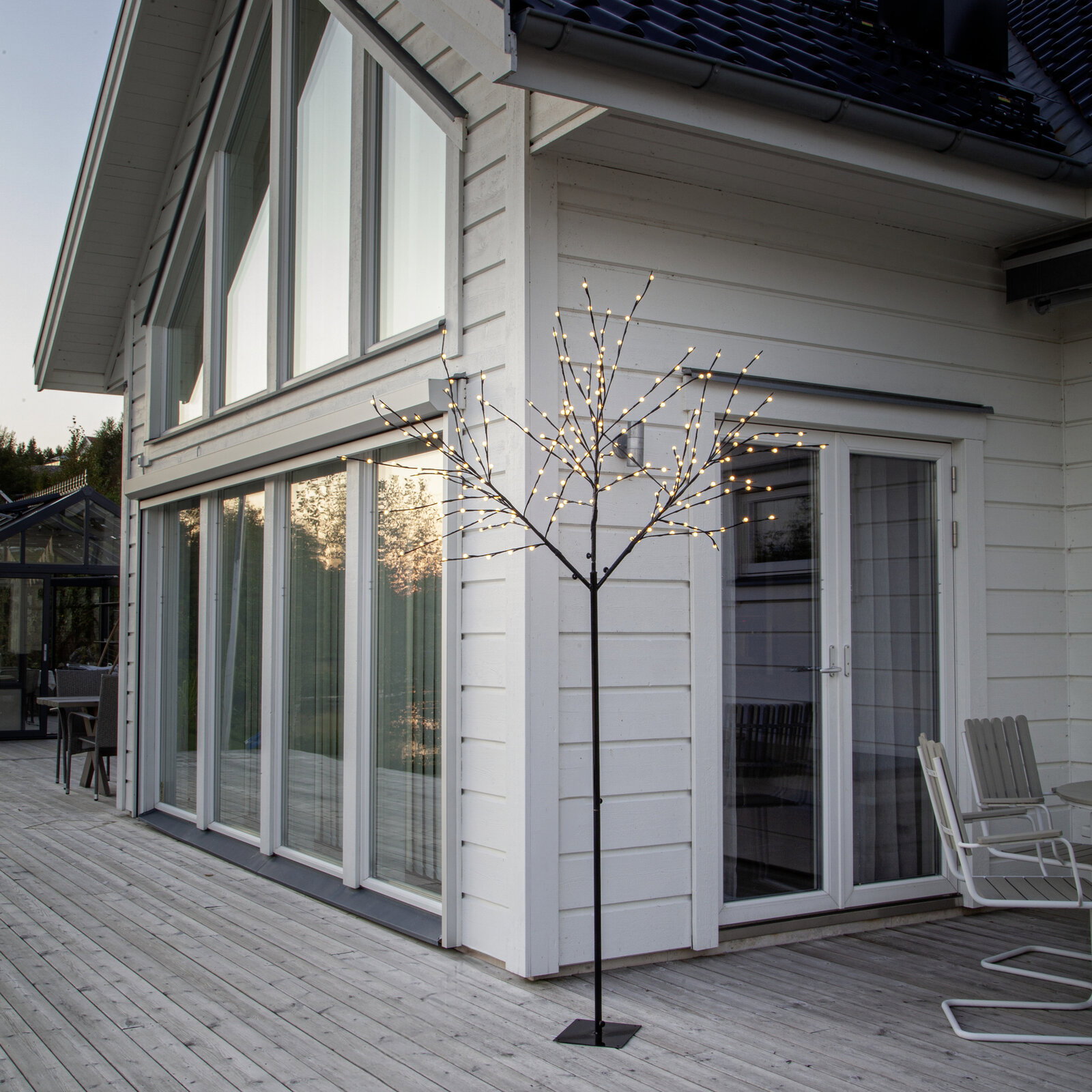 Schwarzer LED-Lichtbaum als dekorative Stehleuchte auf einer Holzterrasse vor einem weißen Haus, warmweiße Lichter, Perspektive: leicht seitliche Außenansicht auf Augenhöhe.