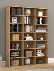 Großes, offenes Bücherregal aus hellem Holz mit vielen quadratischen Fächern, dekoriert mit Büchern, Boxen, Vasen und Pflanzen, frontale Ansicht in einem Wohnraum.