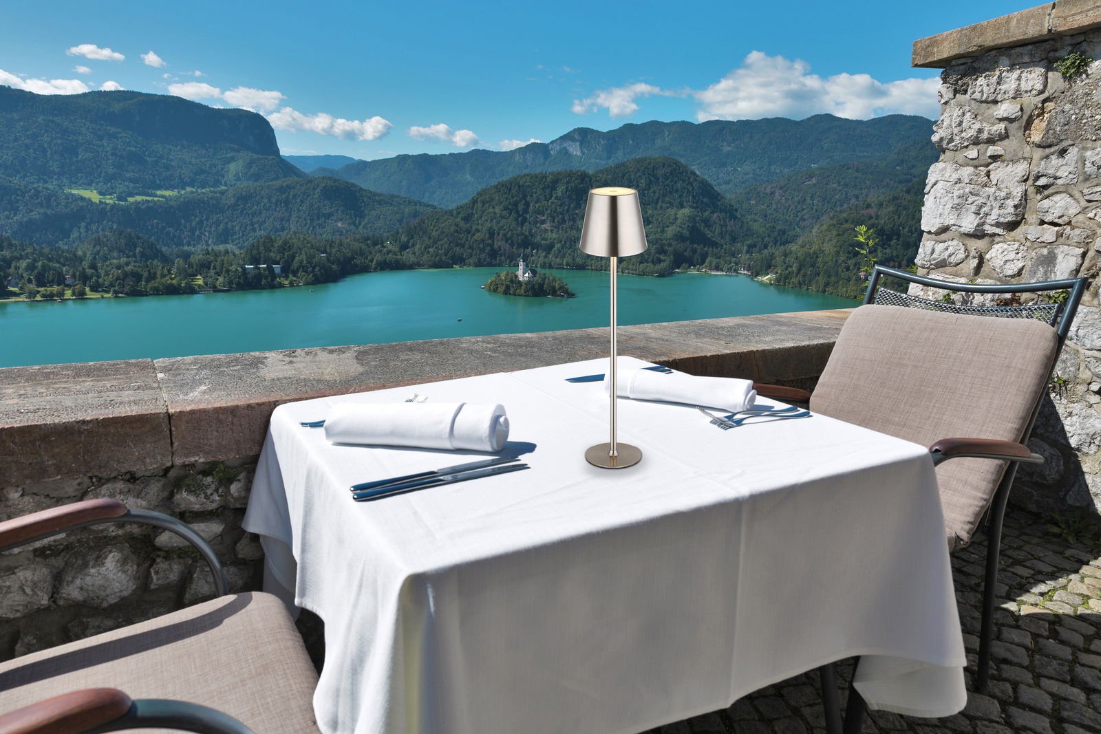 Elegante Tischleuchte VANNIE auf einem gedeckten Tisch mit Blick auf einen malerischen See und Berge im Hintergrund, seitliche Perspektive.