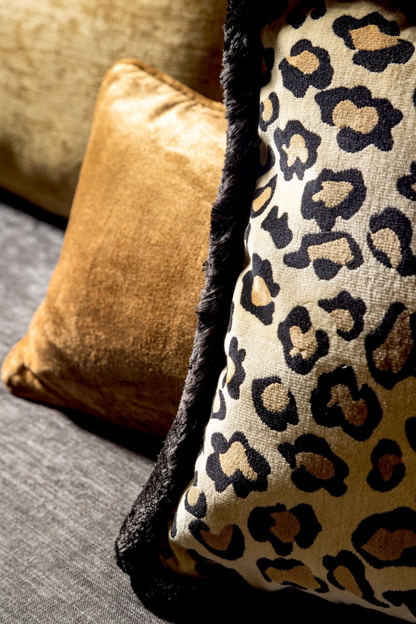 Nahaufnahme von zwei Dekokissen, eines mit Leopardenmuster und schwarzem Rand, das andere in goldener Farbe, auf einem grauen Sofa.