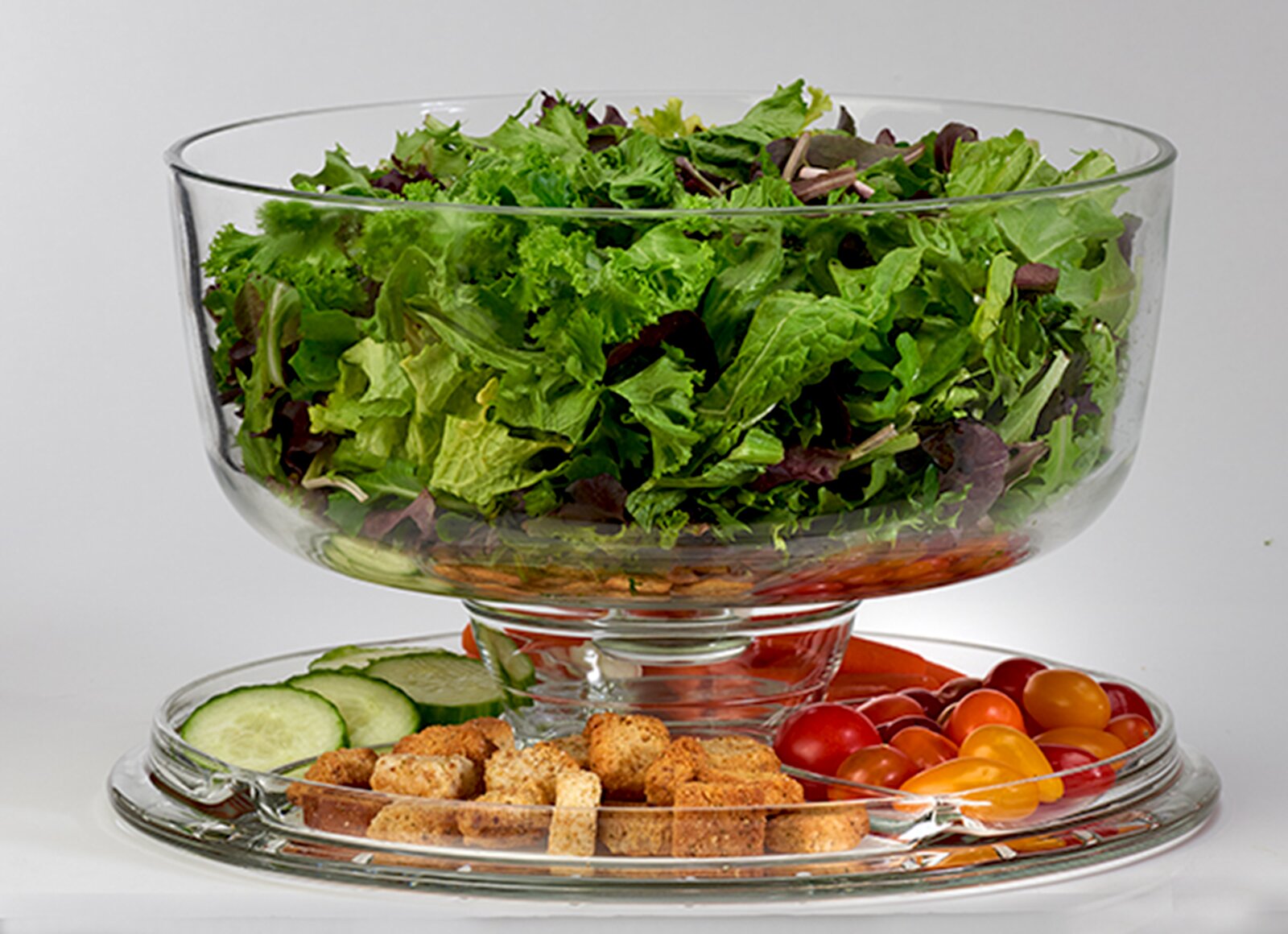 creaTable Servierbutler 2-tlg Transparente Glas-Salatschüssel auf Fuß mit gemischtem Blattsalat, darunter auf einem Glasteller Gurkenscheiben, Croutons und bunte Cherrytomaten; Frontansicht in leicht erhöhter Perspektive.