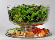 Transparente Glas-Salatschüssel auf Fuß mit gemischtem Blattsalat, darunter auf einem Glasteller Gurkenscheiben, Croutons und bunte Cherrytomaten; Frontansicht in leicht erhöhter Perspektive.