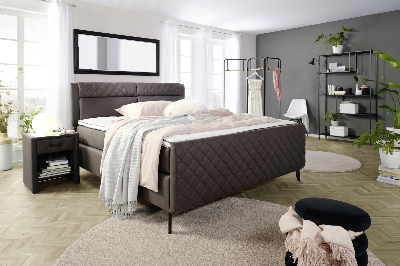 Modernes Schlafzimmer mit einem Boxspringbett in dunklem Stoffbezug, flankiert von einer Nachtkommode. Das Bett ist mit pastellfarbenen Bettwaren dekoriert. Im Hintergrund sind ein Kleiderständer und ein Regal zu sehen. Die Perspektive zeigt das Bett von