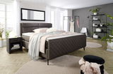 Modernes Schlafzimmer mit einem Boxspringbett in dunklem Stoffbezug, flankiert von einer Nachtkommode. Das Bett ist mit pastellfarbenen Bettwaren dekoriert. Im Hintergrund sind ein Kleiderständer und ein Regal zu sehen. Die Perspektive zeigt das Bett von 