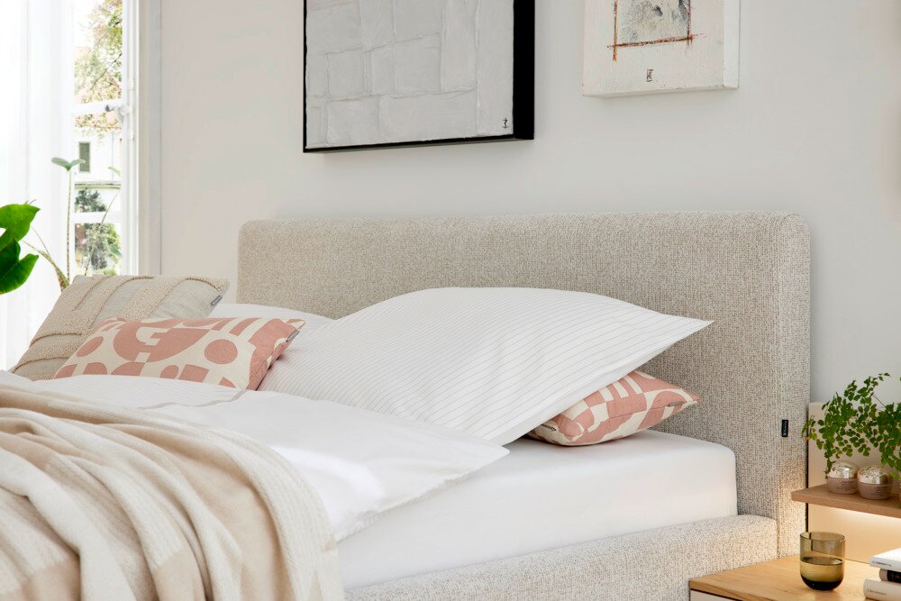 Zijaanzicht van een gestoffeerd bed met een beige hoofdeinde, versierd met witte en gedessineerde kussens in een lichte, moderne slaapkamer.