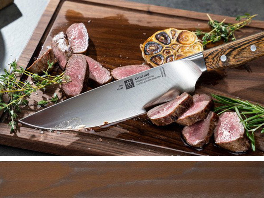 Ein großes Messer liegt auf einem Holzbrett. Daneben sind Fleischstücke, Knoblauch und Kräuter.
