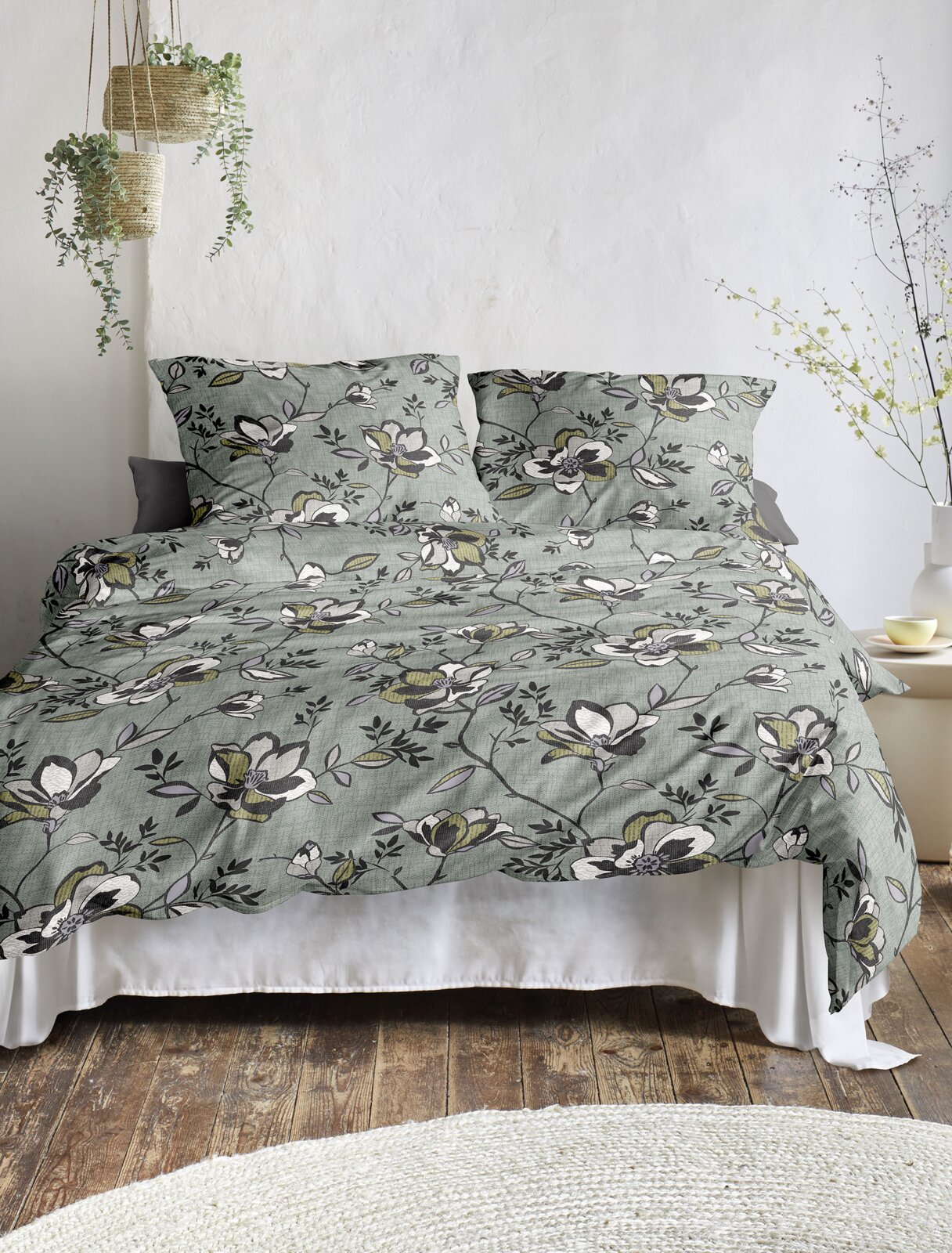 Bettwäsche-Set mit floralem Muster in Grün- und Weißtönen, auf einem Bett in einem hellen Raum mit hängenden Pflanzen, aus der Frontalperspektive.