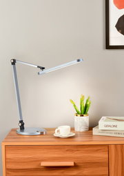 Moderne LED-Schreibtischleuchte in seitlicher Perspektive auf einem Holztisch, neben einer Tasse, Büchern und einer kleinen Pflanze.