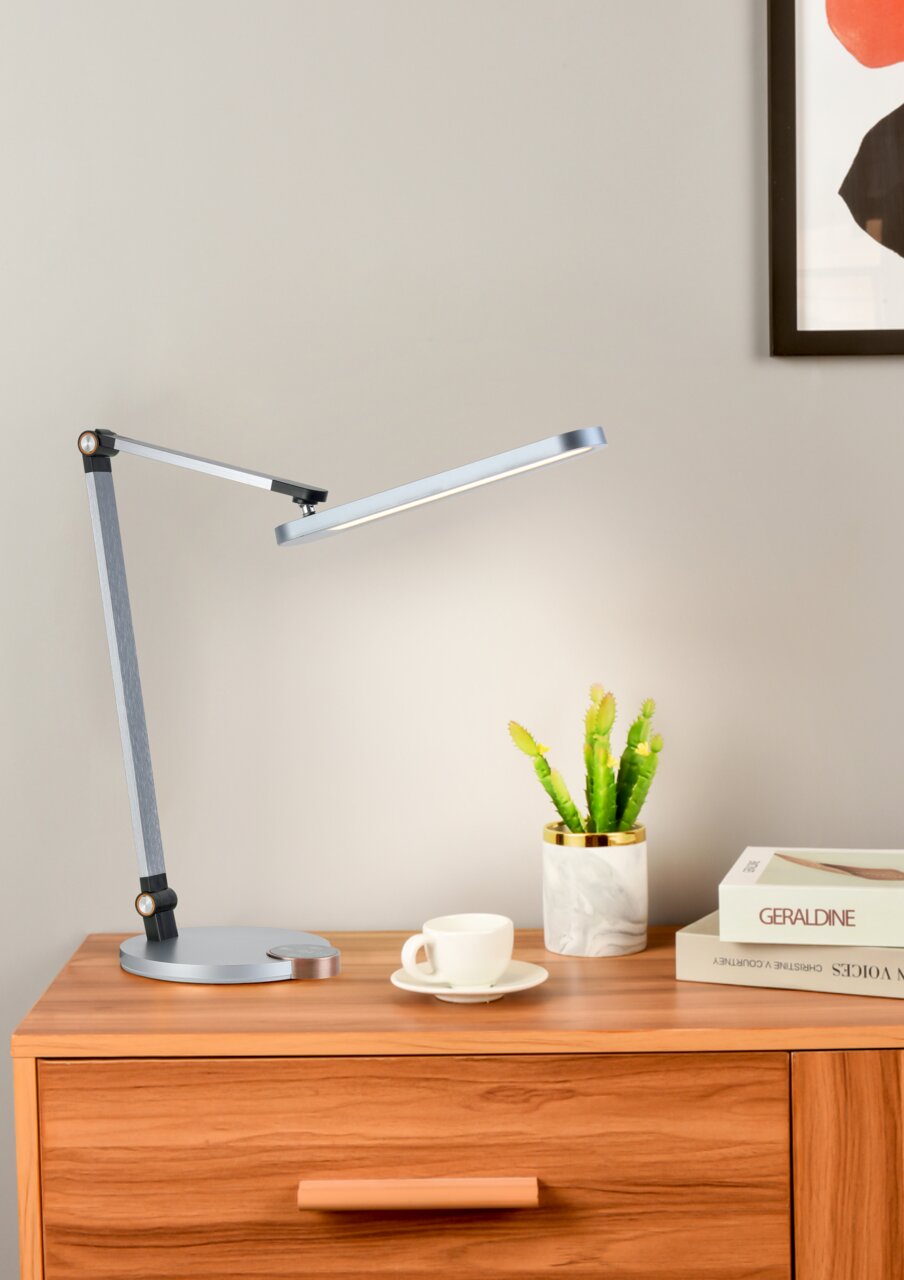 Moderne LED-Schreibtischleuchte in seitlicher Perspektive auf einem Holztisch, neben einer Tasse, Büchern und einer kleinen Pflanze.