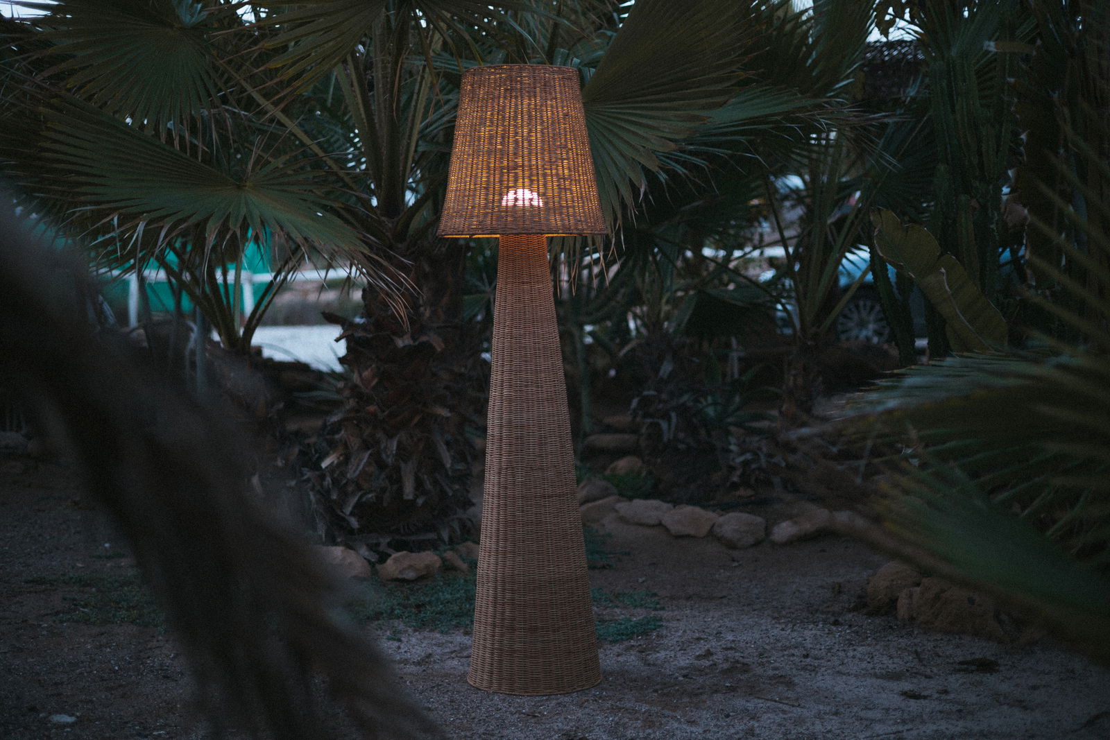 Akku-Outdoor-Stehleuchte aus Rattan in einem Garten bei Dämmerung, seitliche Perspektive