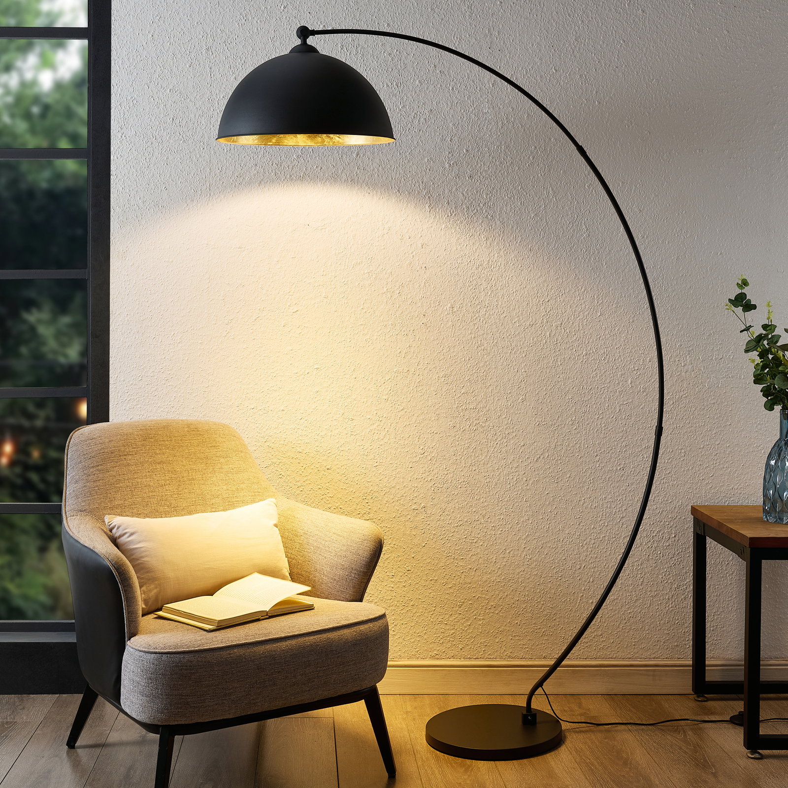 Novel Stehleuchte Moderne Stehleuchte in Bogenform, die von der Seite betrachtet wird und ein gemütliches Licht auf einen Sessel und ein Buch wirft.