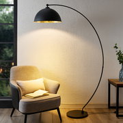 Moderne Stehleuchte in Bogenform, die von der Seite betrachtet wird und ein gemütliches Licht auf einen Sessel und ein Buch wirft.