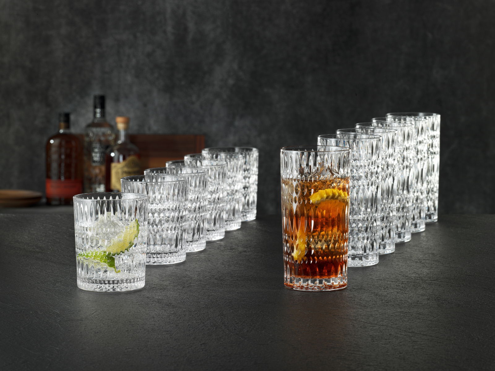 Ein 12-teiliges Becherset im Ethno-Stil, bestehend aus klaren, strukturierten Gläsern, die auf einer dunklen Oberfläche in einer diagonalen Anordnung präsentiert werden. Im Vordergrund sind zwei Gläser mit Getränken und Eiswürfeln zu sehen.