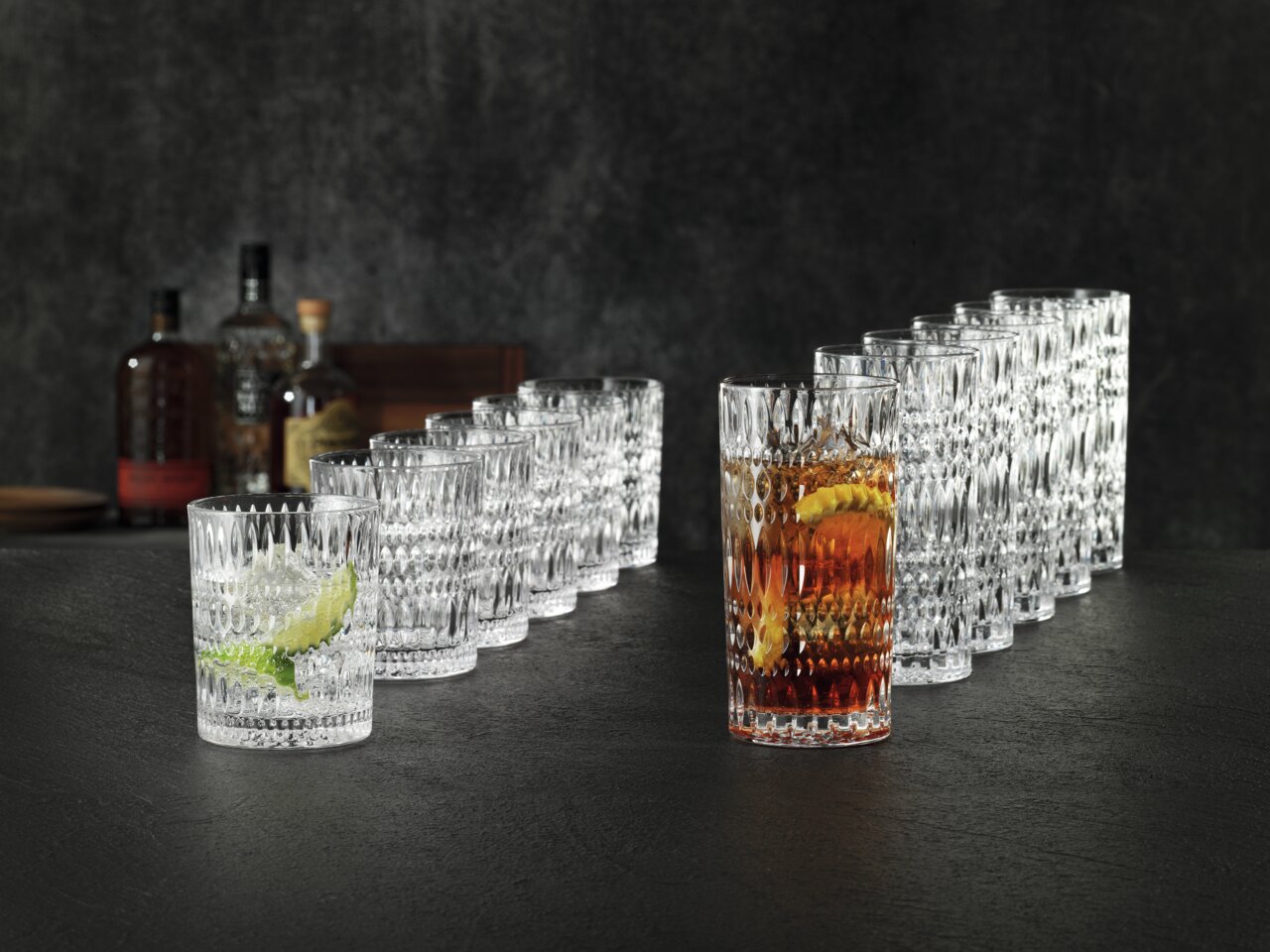 Nachtmann Becherset 12-tlg. ETHNO Ein 12-teiliges Becherset im Ethno-Stil, bestehend aus klaren, strukturierten Gläsern, die auf einer dunklen Oberfläche in einer diagonalen Anordnung präsentiert werden. Im Vordergrund sind zwei Gläser mit Getränken und Eiswürfeln zu sehen.