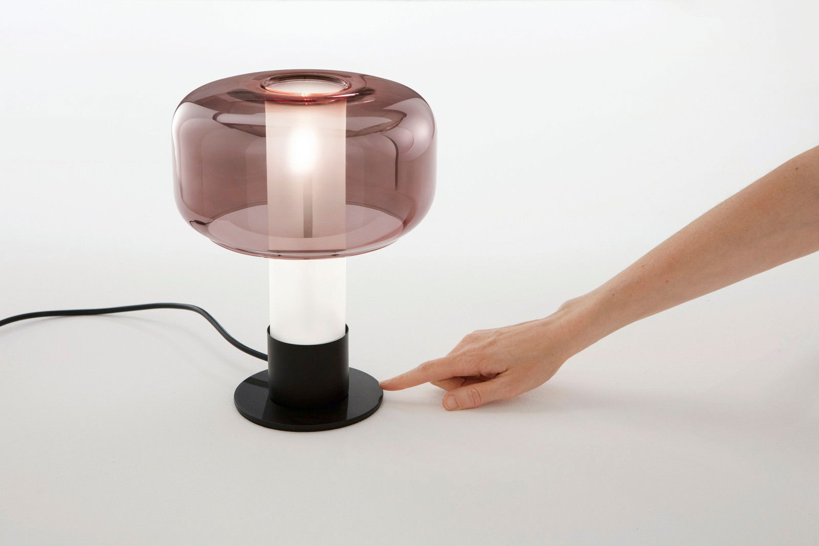 Moderne Tischleuchte mit rauchigem Glas und schwarzem Sockel, seitliche Perspektive, Hand zeigt auf den Sockel.