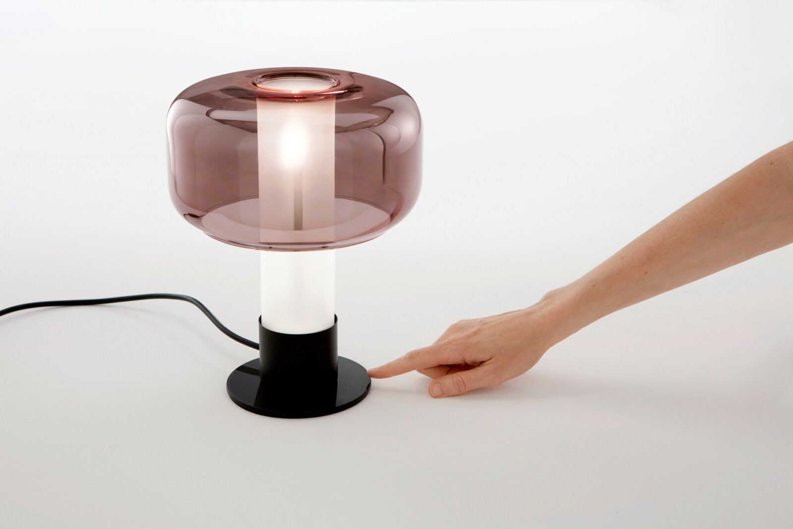 Moderne Tischleuchte mit rauchigem Glas und schwarzem Sockel, seitliche Perspektive, Hand zeigt auf den Sockel.