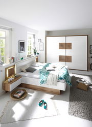 Moderne Schlafzimmeransicht mit einem weißen Bett und einem Schwebetürenschrank in Holz- und Weißtönen, von der Seite betrachtet.