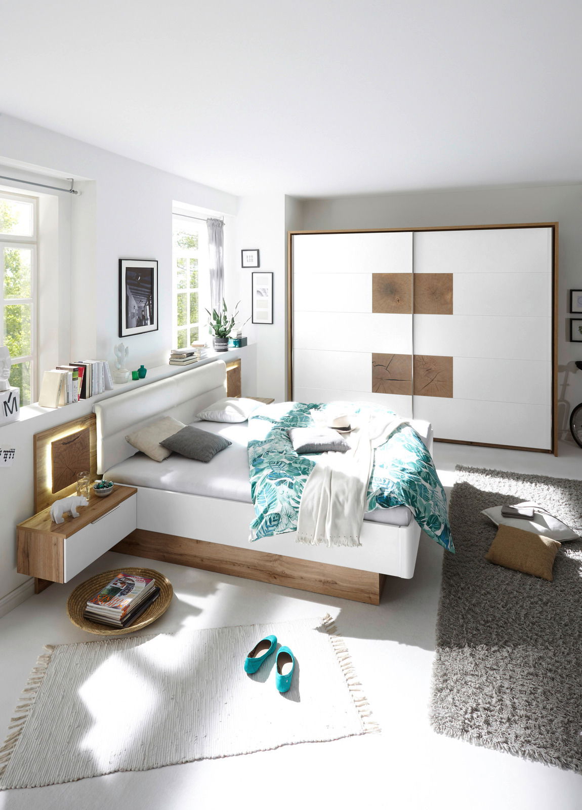 Moderne Schlafzimmeransicht mit einem weißen Bett und einem Schwebetürenschrank in Holz- und Weißtönen, von der Seite betrachtet.