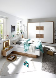 Moderne Schlafzimmeransicht mit einem weißen Bett und einem Schwebetürenschrank in Holz- und Weißtönen, von der Seite betrachtet.