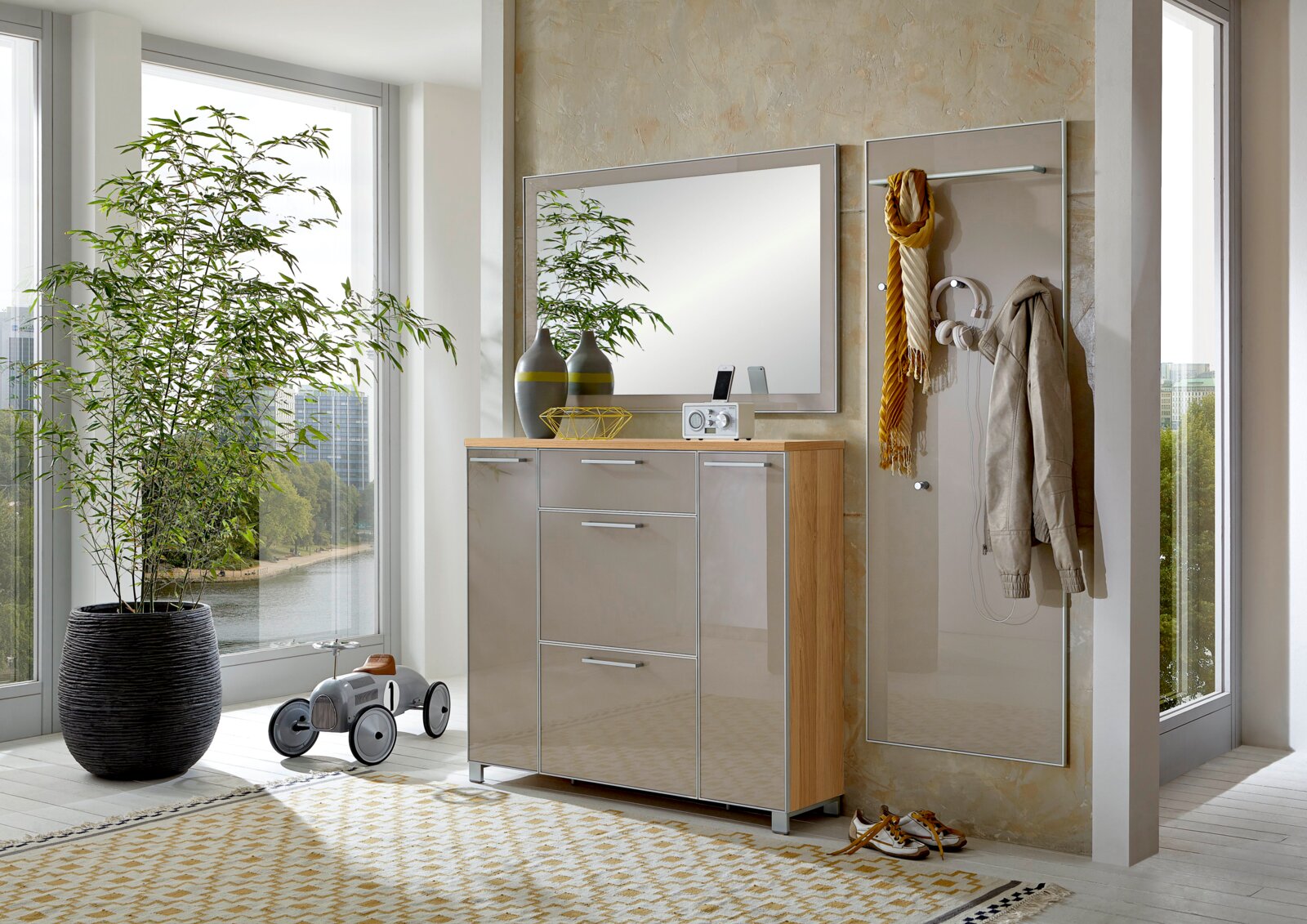 Modernes Garderoben-Set in einem hellen Raum mit großen Fenstern, bestehend aus einem Spiegel, einem Schrank mit Hochglanzfronten und einer Wandgarderobe mit Haken. Perspektive von vorne.