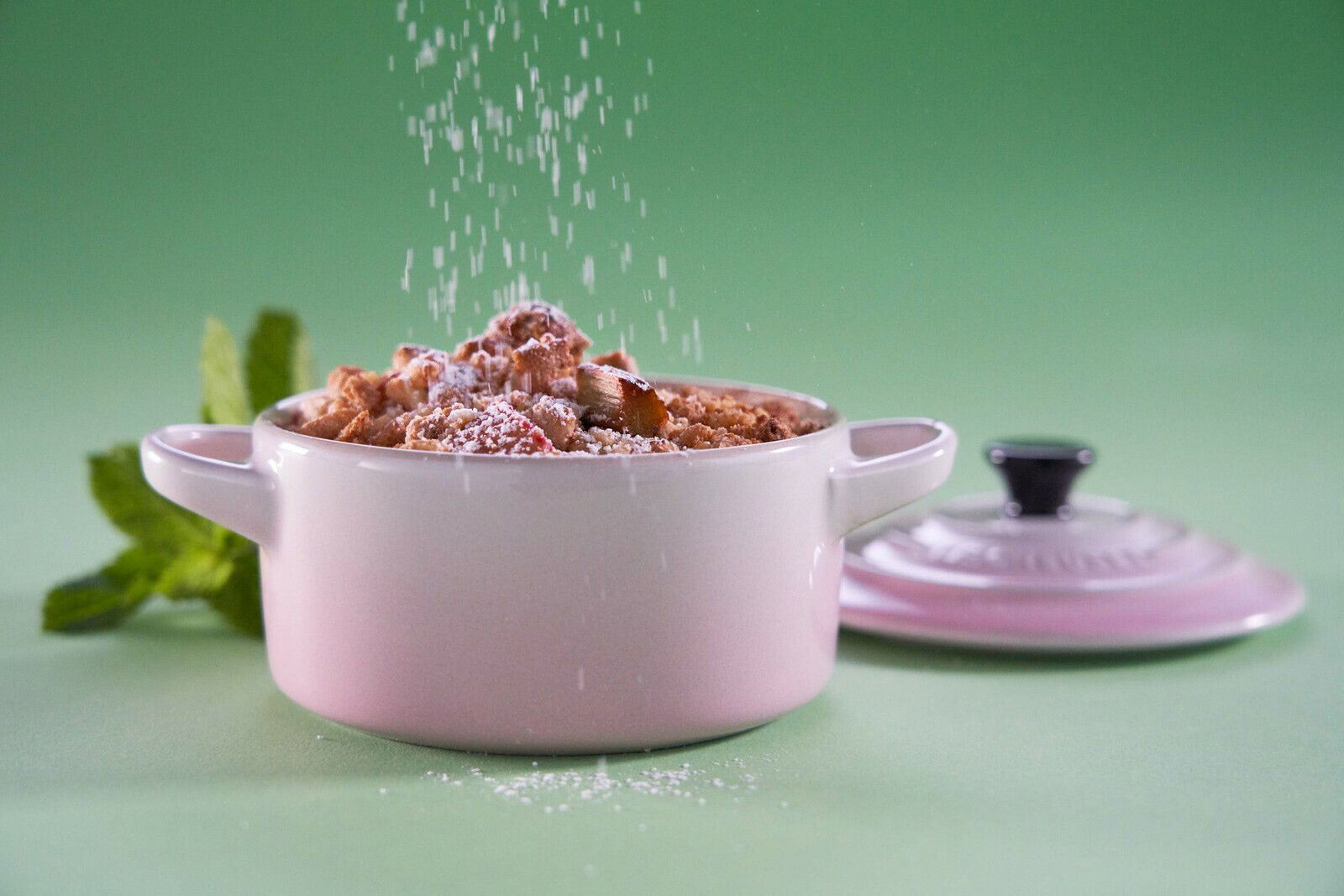 LE CREUSET Mini Cocotte 10cm Shell Pink Mini-Cocotte in Muschelrosa mit Streuselkuchen, von vorne fotografiert, mit Puderzucker bestreut, auf grünem Hintergrund.