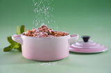 Mini-Cocotte in Muschelrosa mit Streuselkuchen, von vorne fotografiert, mit Puderzucker bestreut, auf grünem Hintergrund.