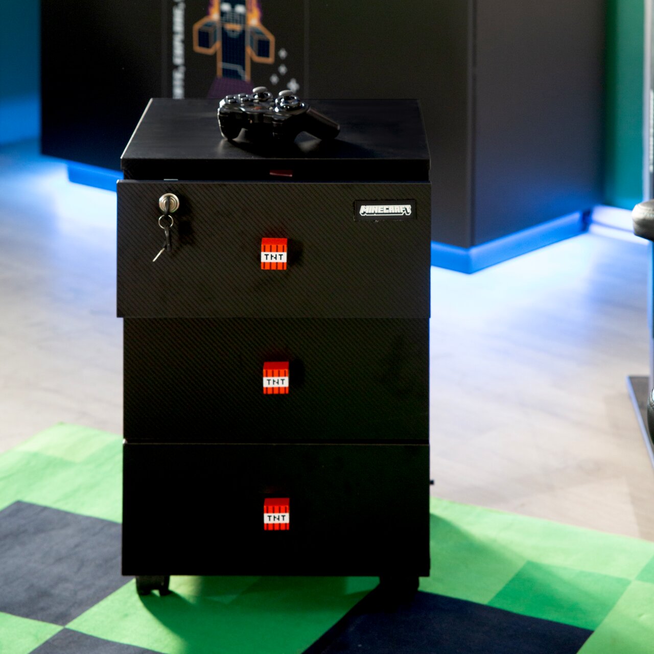 Zwarte, verrijdbare Minecraft-ladekast met drie lades, TNT-logo’s en een slot met sleutel; bovenop ligt een gamecontroller; vooraanzicht licht van bovenaf.