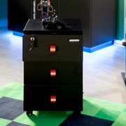 Zwarte, verrijdbare Minecraft-ladekast met drie lades, TNT-logo’s en een slot met sleutel; bovenop ligt een gamecontroller; vooraanzicht licht van bovenaf.