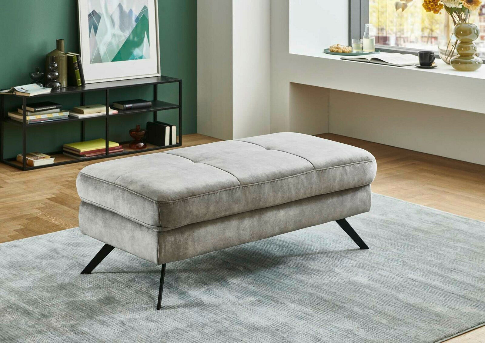 Grauer XXL-Hocker mit schwarzen Metallbeinen in einem modernen Wohnzimmer, seitliche Perspektive.