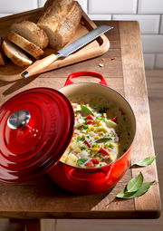 Roter ovaler Bräter mit geöffnetem Deckel, gefüllt mit einem bunten Eintopf, auf einem Holztisch neben geschnittenem Brot. Perspektive von oben.