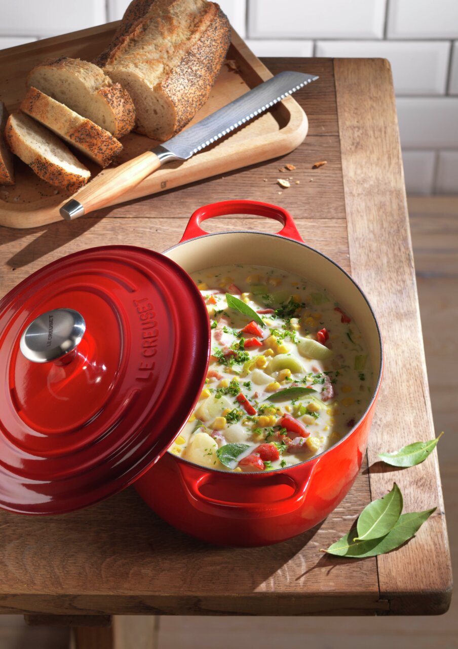 LE CREUSET Bräter Oval 29cm Kirschrot SIGNATURE Roter ovaler Bräter mit geöffnetem Deckel, gefüllt mit einem bunten Eintopf, auf einem Holztisch neben geschnittenem Brot. Perspektive von oben.