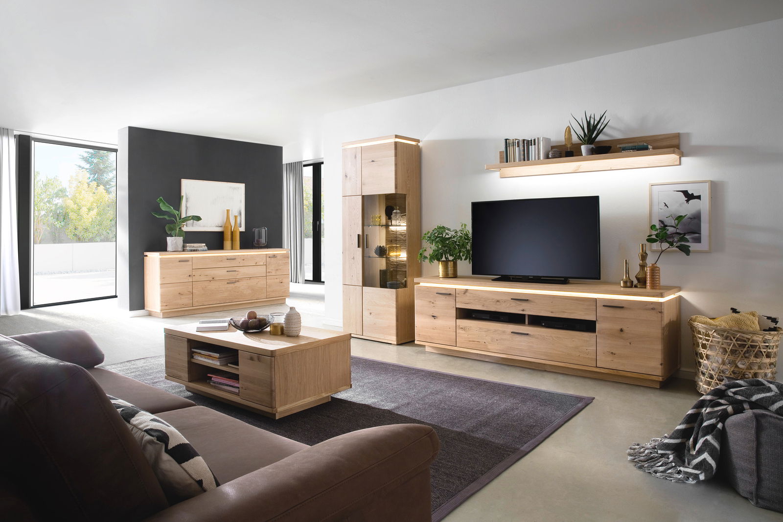 Wohnzimmer mit moderner Holzmöbelkombination, bestehend aus Sideboard, Vitrine, TV-Element und Wandboard, aus der Perspektive eines Sofas betrachtet.
