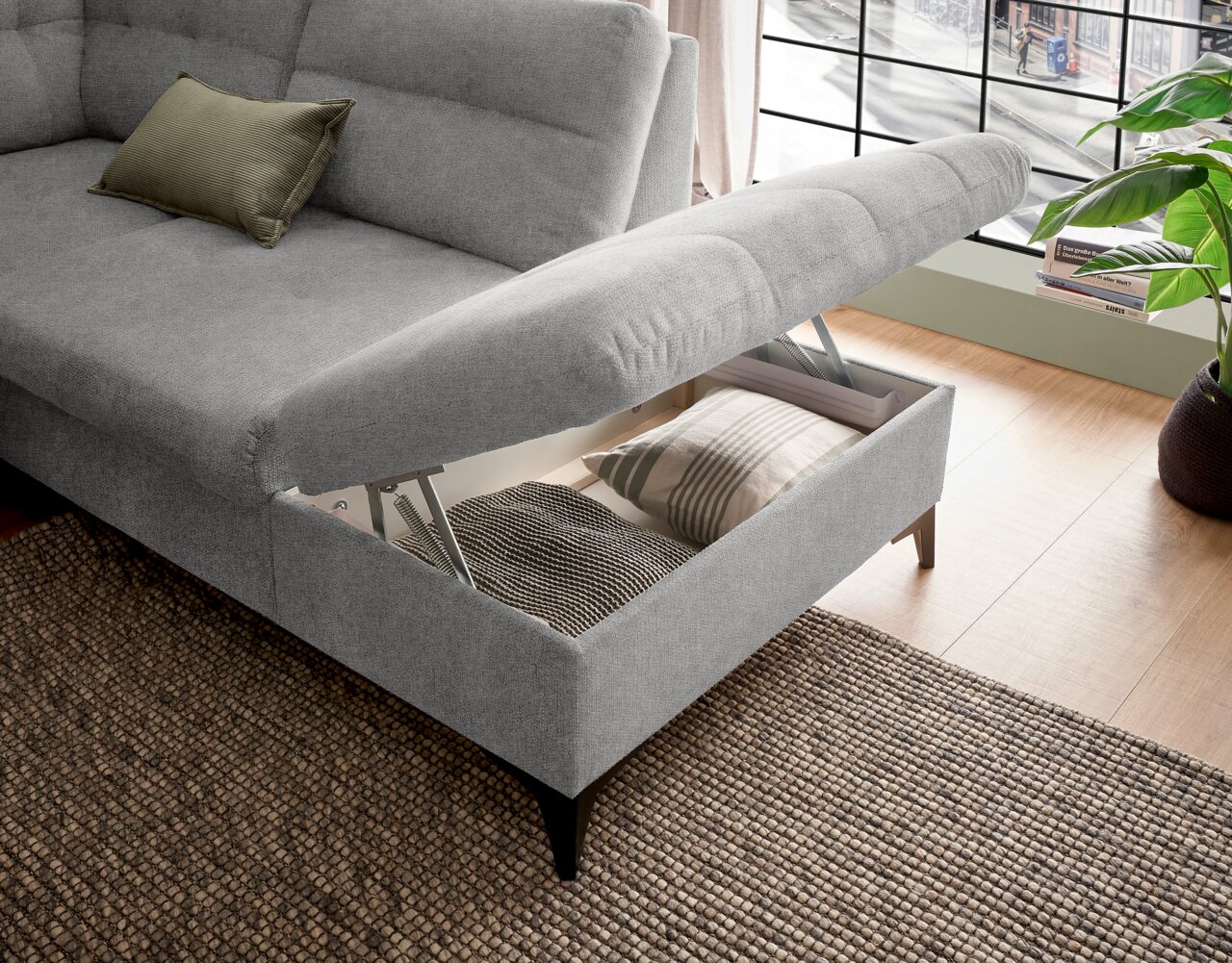 Graues Ecksofa mit Stauraum, geöffnet, von schräg oben fotografiert. Ein Kissen liegt auf der Sitzfläche, und im Stauraum sind weitere Kissen sichtbar.