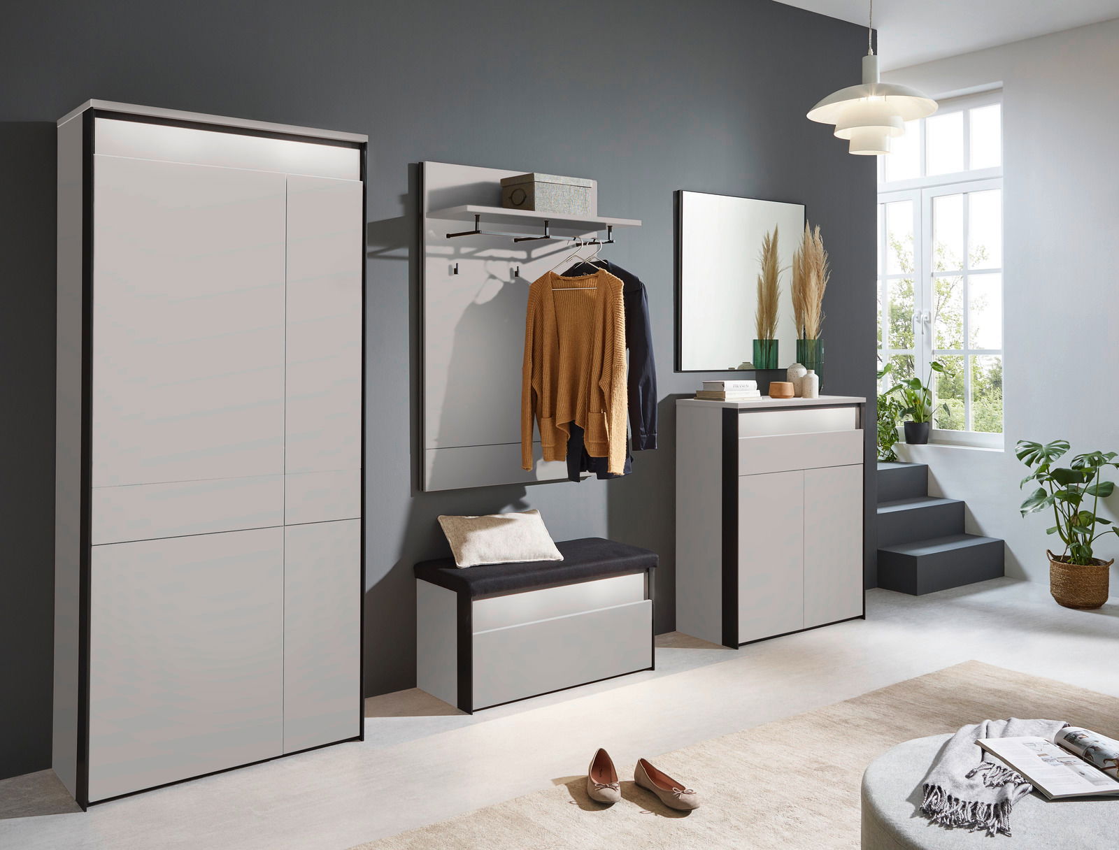 Voleo Schuhschrank ZAYA Moderne Garderobeneinrichtung in einem hellen Raum mit großem Fenster, bestehend aus einem XL-Garderobenschrank, einer Garderobenbank mit Kissen, einem Garderobenpaneel mit Kleiderstange und einem Sideboard, seitliche Perspektive.