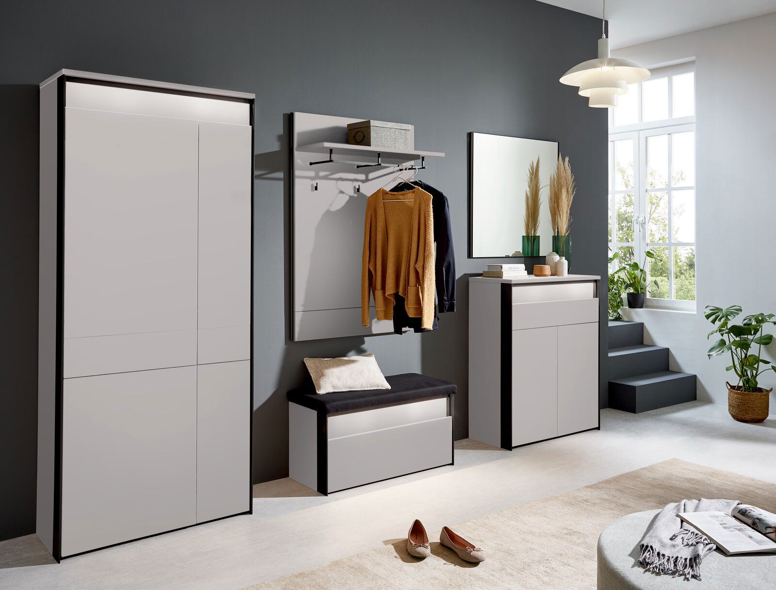 Voleo Garderobenbank ZAYA Moderne Garderobeneinrichtung in einem hellen Raum mit großem Fenster, bestehend aus einem XL-Garderobenschrank, einer Garderobenbank mit Kissen, einem Garderobenpaneel mit Kleiderstange und einem Sideboard, seitliche Perspektive.