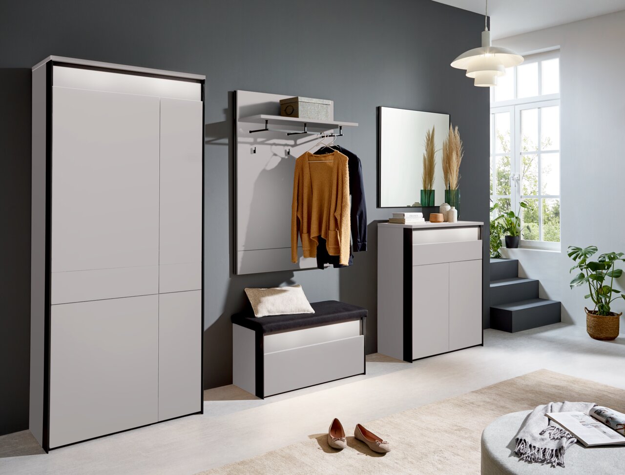 Voleo Schuhschrank ZAYA Moderne Garderobeneinrichtung in einem hellen Raum mit großem Fenster, bestehend aus einem XL-Garderobenschrank, einer Garderobenbank mit Kissen, einem Garderobenpaneel mit Kleiderstange und einem Sideboard, seitliche Perspektive.