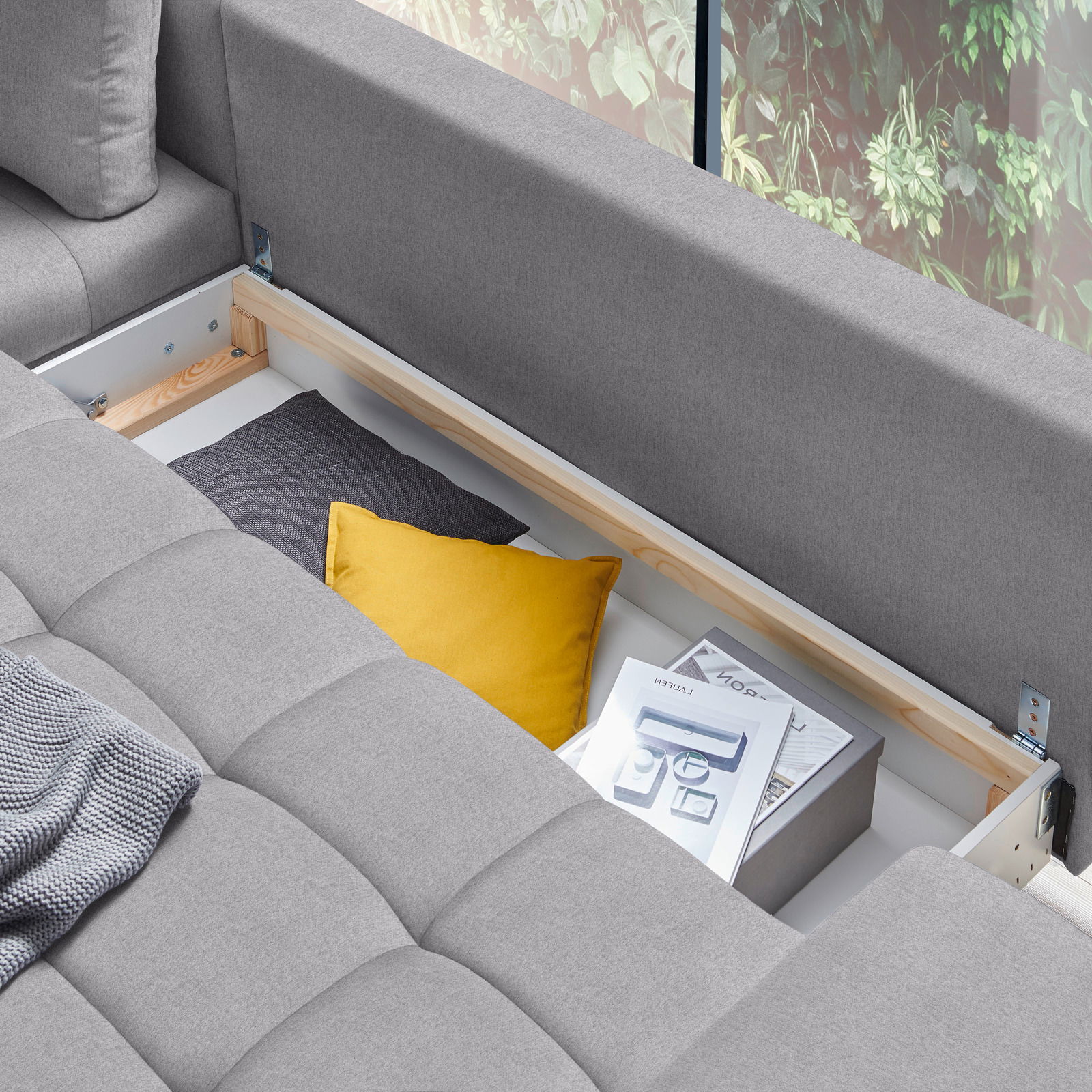 Graue Rundecke Ottli mit geöffneter Stauraumfunktion, gelbem Kissen und Zeitschriften, von oben betrachtet.