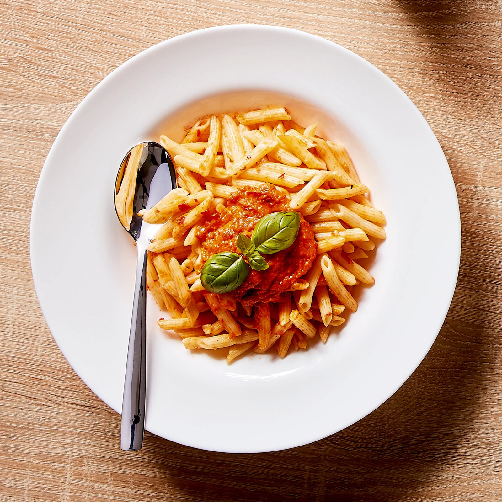 Draufsicht auf einen weißen Teller mit Penne-Pasta in Tomatensauce, garniert mit frischem Basilikum, daneben ein Löffel auf Holzuntergrund