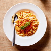Draufsicht auf einen weißen Teller mit Penne-Pasta in Tomatensauce, garniert mit frischem Basilikum, daneben ein Löffel auf Holzuntergrund