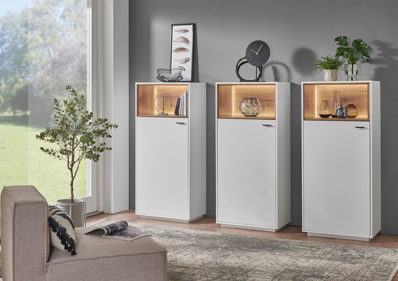 Zurbrüggen Hoge vitrinekast CORUNA Drei moderne Highboard-Vitrinen in Weiß mit beleuchteten Holzfächern, dekoriert mit Pflanzen und Kunstobjekten, in einem stilvollen Wohnzimmer. Perspektive: Frontalansicht.
