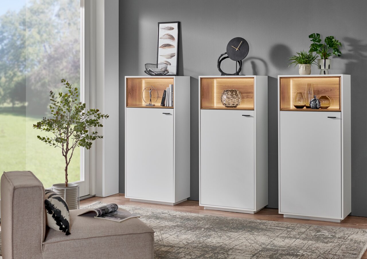 Zurbrüggen Highboard-Vitrine CORUNA Drei moderne Highboard-Vitrinen in Weiß mit beleuchteten Holzfächern, dekoriert mit Pflanzen und Kunstobjekten, in einem stilvollen Wohnzimmer. Perspektive: Frontalansicht.