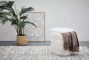 Hocker mit Stauraum in weißem, flauschigem Design, von der Seite betrachtet, mit einer braunen Decke darauf, neben einer Pflanze und einem Bild.