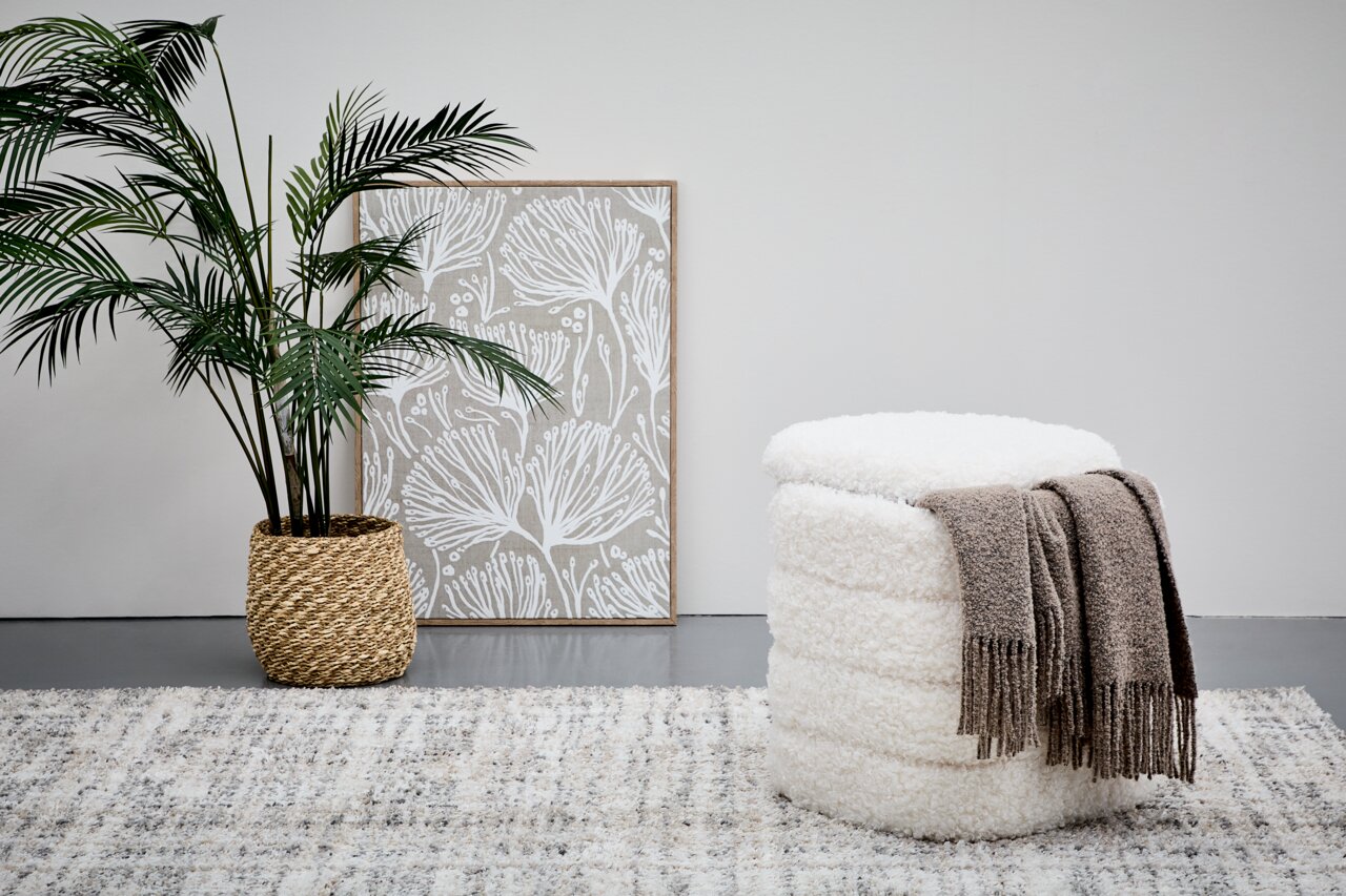 Hocker mit Stauraum in weißem, flauschigem Design, von der Seite betrachtet, mit einer braunen Decke darauf, neben einer Pflanze und einem Bild.