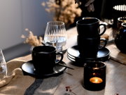 Schwarzes Kaffeetassen- und Untertassen-Set auf rustikalem Holztisch, eine Tasse im Vordergrund und zwei gestapelte Tassen rechts, seitliche Perspektive mit warmem Kerzenlicht und Glas im Hintergrund.